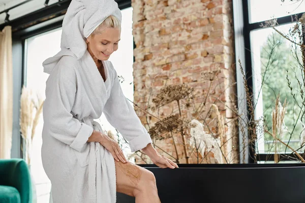 Heureuse femme d'âge moyen en peignoir et avec serviette sur la tête appliquer gommage sur la jambe près de la baignoire — Photo de stock