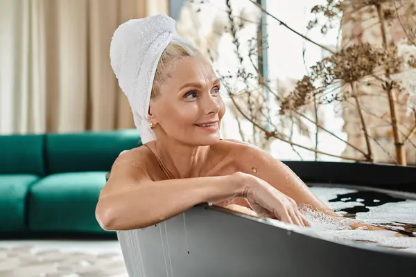 Задоволена жінка середнього віку з білим рушником на голові приймає ванну в сучасній квартирі, релакс — стокове фото