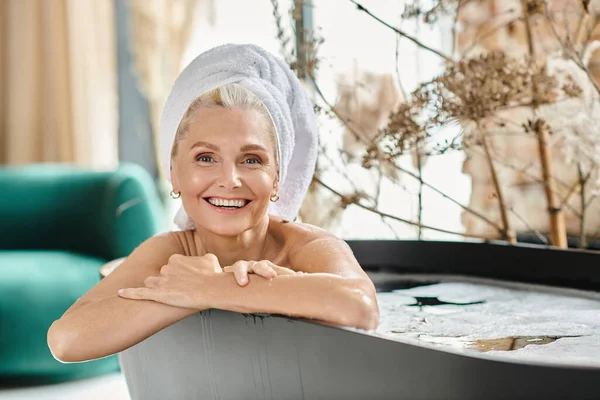 Heureux femme d'âge moyen avec serviette blanche sur la tête prenant bain dans un appartement moderne, spa à la maison — Photo de stock