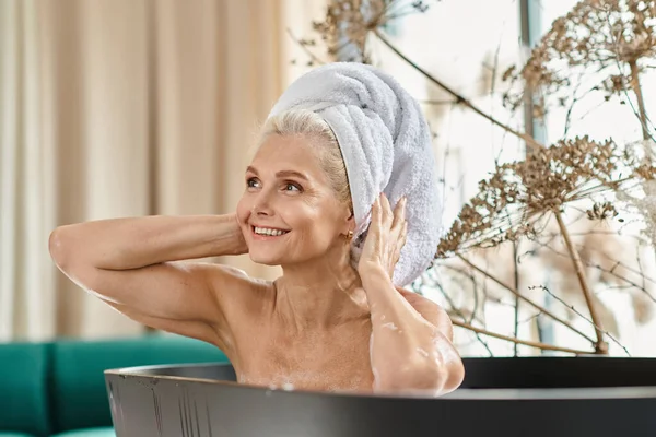 Gaie femme d'âge moyen avec serviette blanche sur la tête prenant bain dans un appartement moderne, spa à la maison — Photo de stock