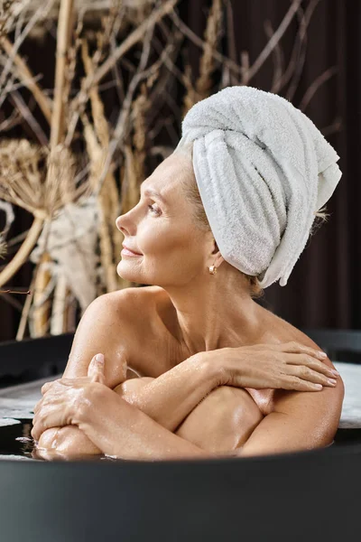 Приваблива жінка середнього віку з білим рушником на голові приймає ванну в сучасній квартирі, спа-день — стокове фото