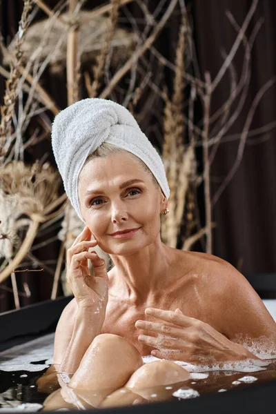 Красива жінка середнього віку з білим рушником на голові приймає ванну в сучасній квартирі, спа-день — стокове фото