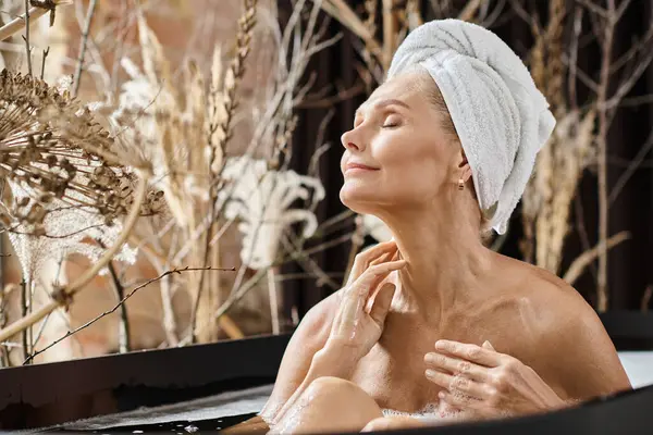 Довольная женщина среднего возраста с полотенцем на голове и закрытыми глазами принимая ванну в современной квартире — стоковое фото
