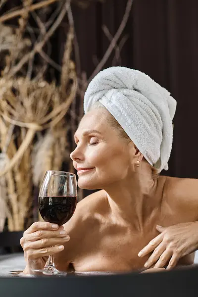 Pleased middle aged woman with towel on head holding glass of red wine while taking bath at home — Stock Photo