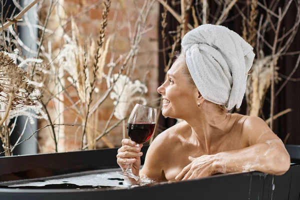 Positive middle aged woman with towel on head holding glass of red wine while taking bath at home — Stock Photo