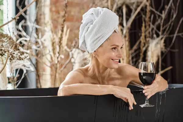 Calm and happy middle aged woman with towel on head holding glass of red wine while taking bath — Stock Photo