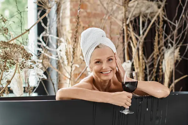 Розслаблена і щаслива жінка середнього віку з рушником на голові тримає келих червоного вина під час прийняття ванни — стокове фото