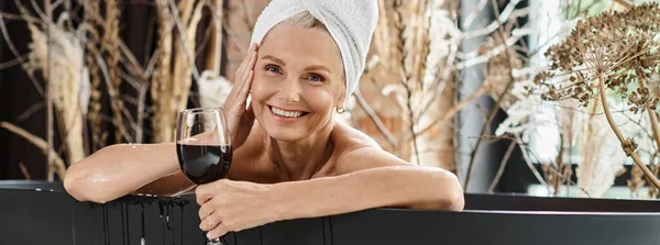 Bannière de heureuse femme d'âge moyen avec serviette sur la tête tenant un verre de vin rouge tout en prenant un bain — Photo de stock