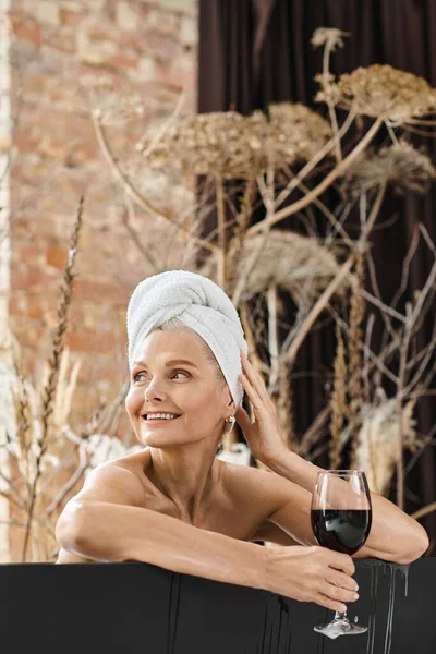 Relaxado e alegre mulher de meia-idade com toalha na cabeça segurando copo de vinho tinto enquanto toma banho — Fotografia de Stock
