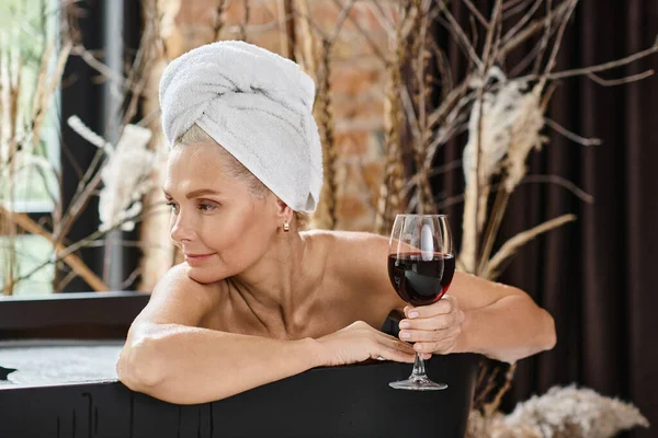 Розслаблена жінка середнього віку з білим рушником на голові тримає келих червоного вина і приймає ванну — стокове фото
