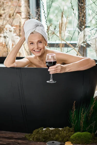 Сяюча жінка середнього віку з білим рушником на голові тримає келих червоного вина і приймає ванну — стокове фото