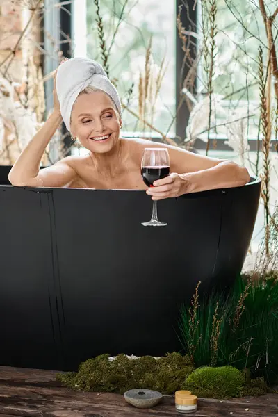 Усміхнена жінка середнього віку з білим рушником на голові тримає келих червоного вина і приймає ванну — стокове фото