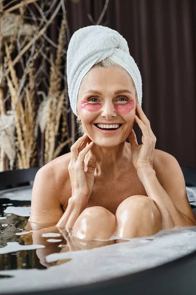 Cheerful middle aged woman with white towel on head and under eye patches taking bath at home — Stock Photo