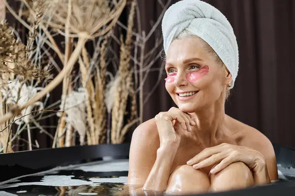 Joyeuse femme d'âge moyen avec serviette blanche sur la tête et sous les yeux patchs prendre un bain à la maison — Photo de stock