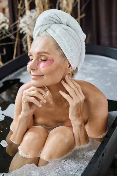 Mujer de mediana edad complacida con toalla blanca en la cabeza y bajo los parches de los ojos tomando baño en casa - foto de stock