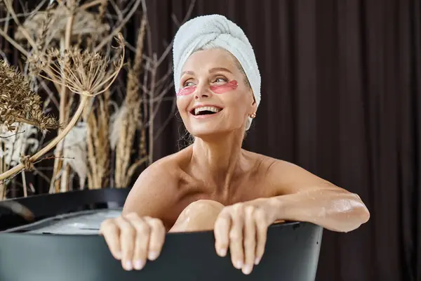 Mujer de mediana edad excitada con toalla blanca en la cabeza y bajo los parches de los ojos tomando baño en casa - foto de stock