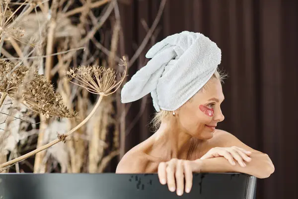 Jolie femme d'âge moyen avec serviette blanche sur la tête et sous les yeux patchs prendre un bain à la maison — Photo de stock