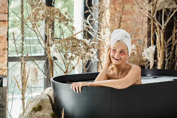 Joyous middle aged woman with towel on head and eye patches taking bath in modern apartment — Stock Photo