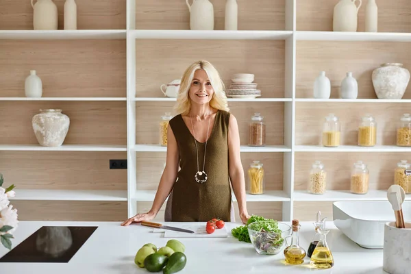 Happy blonde middle aged woman standing next to countertop with fresh ingredients and electric stove — Stock Photo