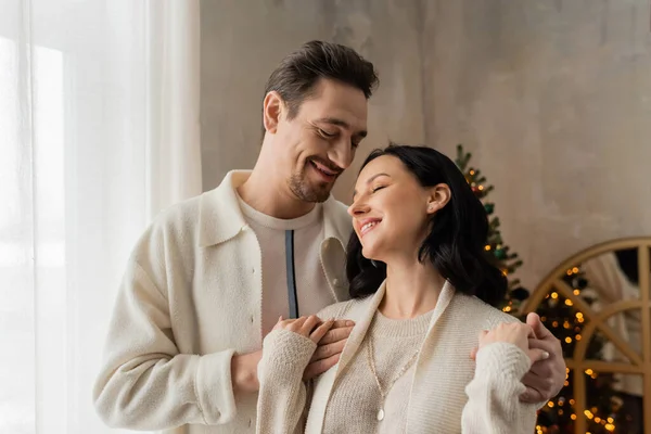 Homem alegre com barba abraçando esposa feliz no aconchegante casa desgaste perto borrada árvore de Natal no pano de fundo — Fotografia de Stock