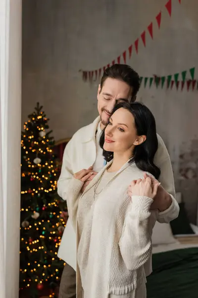 Homme joyeux avec les yeux fermés étreignant femme heureuse dans un vêtement de maison confortable près de l'arbre de Noël flou — Photo de stock