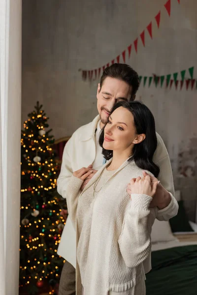 Joyful man with closed eyes hugging happy wife in cozy home wear near blurred Christmas tree — Stock Photo