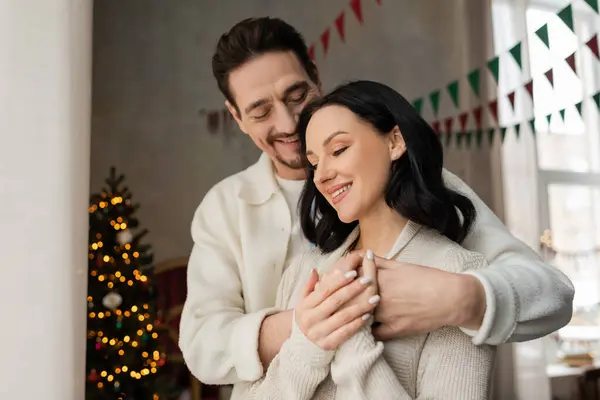 Alegre hombre abrazando esposa en acogedora casa desgaste cerca borrosa decorado árbol de Navidad, vacaciones de invierno - foto de stock