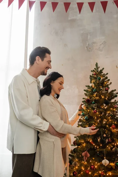 Happily married couple in cozy home wear decorating Christmas tree in modern apartment, festive day — Stock Photo