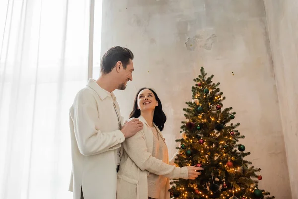 Happily married couple in soft home wear decorating Christmas tree in modern apartment, holiday — Stock Photo