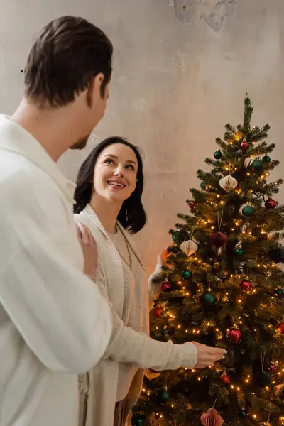 Mujer feliz mirando al marido mientras decoran el árbol de Navidad juntos en casa, vacaciones de invierno - foto de stock