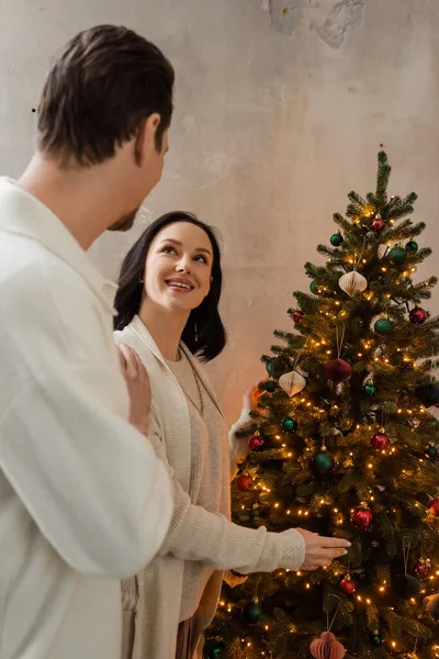 Happy woman looking at husband while decorating Christmas tree together at home, winter holidays — Stock Photo