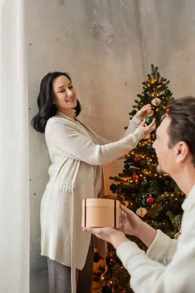 Mulher feliz em casa desgaste segurando bugiganga ao decorar árvore de Natal perto do marido com presente — Fotografia de Stock