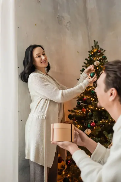 Happy woman in home wear holding bauble while decorating Christmas tree near husband with present — Stock Photo
