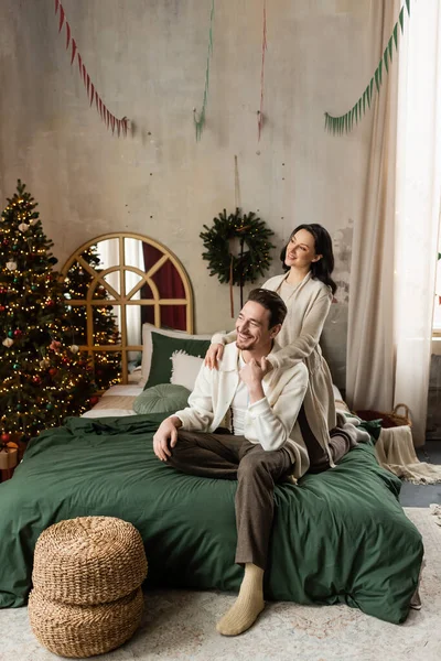 Heureux couple marié étreignant et assis sur le lit près décoré arbre de Noël dans un appartement moderne — Photo de stock