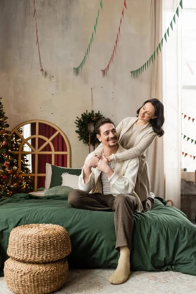 Joyeux couple marié étreignant et assis sur le lit près décoré arbre de Noël dans un appartement moderne — Photo de stock