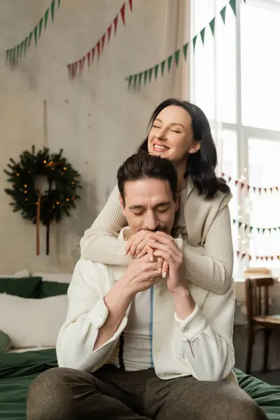 Hombre cariñoso sentado en la cama y besar las manos de la esposa feliz cerca de la corona de Navidad en apartamento moderno - foto de stock