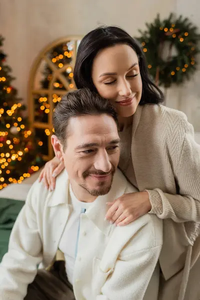 Радостная женщина обнимает бородатого мужа возле размытой елки в современной квартире, довольство — стоковое фото