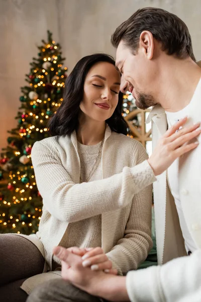 Happy woman with closed eyes sitting with husband near blurred Christmas tree, season of joy — Stock Photo