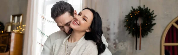Husband embracing joyful wife and smiling together near blurred Christmas wreath on wall, banner — Stock Photo