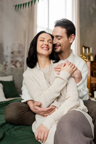 Happy husband embracing joyful wife and sitting together on bed on Christmas morning, winter holiday — Stock Photo