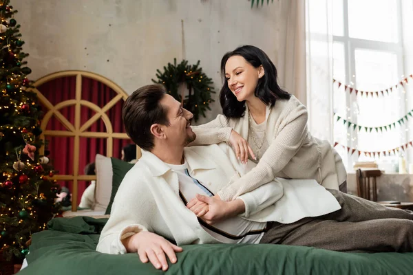 Husband embracing cheerful wife and lying on bed near decorated Christmas tree and wreath on wall — Stock Photo