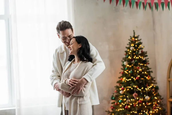 Couple joyeux et marié à la maison porter câlins et debout ensemble près de l'arbre de Noël flou — Photo de stock