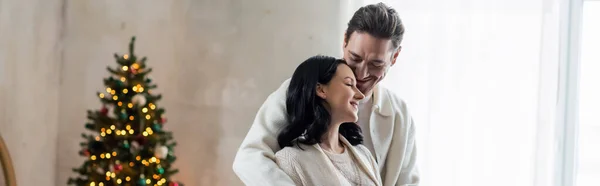 Joyful married couple in home wear hugging and standing together near blurred Christmas tree, banner — Stock Photo