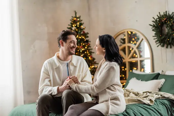 Joyeux mari et femme à la maison portent assis sur le lit et tenant la main près de l'arbre de Noël flou — Photo de stock