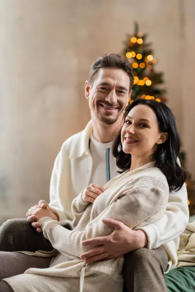 Уютная жизнь, радостная супружеская пара обнимающая друг друга возле размытых рождественских огней на заднем плане — стоковое фото