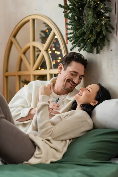 Весёлый мужчина проводит время вместе с женой и лежит на кровати возле рождественского венка на стене — стоковое фото