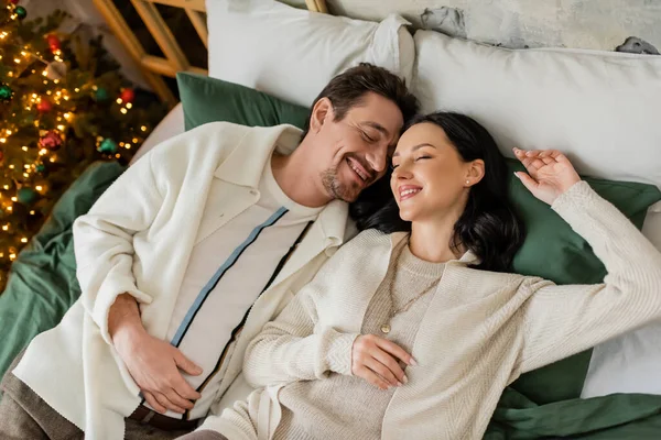 Vista superior de la feliz pareja casada pasar una mañana acogedora en la cama cerca del borroso árbol de Navidad - foto de stock