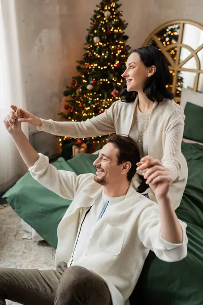 Brünette Frau hält Händchen ihres Mannes und verbringt Zeit zusammen im Schlafzimmer in der Nähe des Weihnachtsbaums — Stockfoto