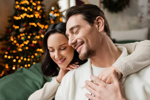Jahreszeit der Freude, glückliche Frau umarmt Ehemann neben geschmücktem Weihnachtsbaum im modernen Schlafzimmer — Stockfoto