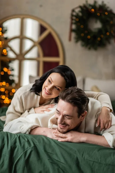 Retrato de la alegre pareja casada mirando hacia otro lado y acostados juntos en la cama cerca del árbol de Navidad - foto de stock